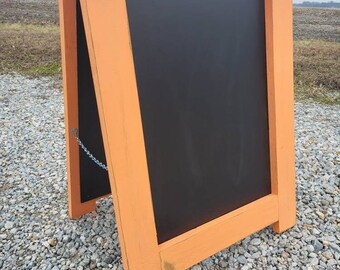 May include: Orange wooden A-frame chalkboard sign with a black chalkboard surface. The sign is standing on a gravel surface.