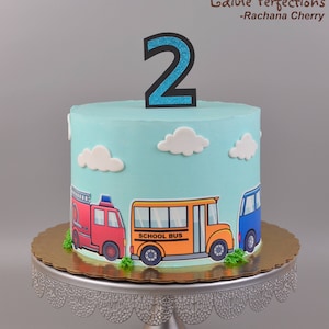 May include: A blue and white birthday cake with a number 2 topper. The cake is decorated with a fire truck, school bus, and a blue car. The cake is on a silver cake stand with a lace design.