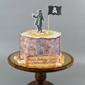 May include: A pirate themed birthday cake with a fondant pirate figure, a pirate flag, and a message that says "Happy Birthday Oviya". The cake is decorated with a map design and is on a wooden cake stand.