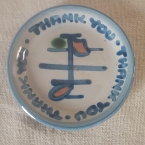 Puede incluir: Un plato pequeño y redondo de cerámica con un borde azul y las palabras "THANK YOU" en escritura azul alrededor del borde. El centro presenta un diseño abstracto azul y naranja con un punto verde.
