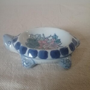 以下が含まれることがあります： 白地にピンクと緑のアクセントの花柄が施された、陶器製のカメ型皿。カメの体は水色で、縁と足に濃い青色のアクセントが付いています。装飾品です。
