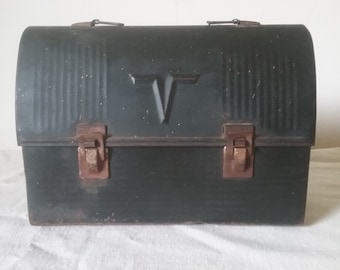 May include: A vintage metal lunchbox with a black finish and a raised letter "V" on the front. The lunchbox has two latches on the front and a handle on the top.