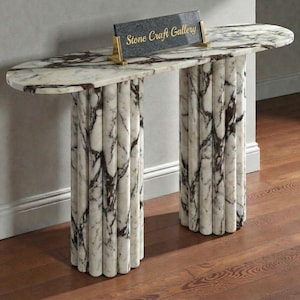 Puede incluir: Una consola de mármol con una tapa con estampado blanco y marrón y dos patas cilíndricas estriadas. La mesa tiene forma semiovalada y presenta un pequeño letrero que dice "Stone Craft Gallery".