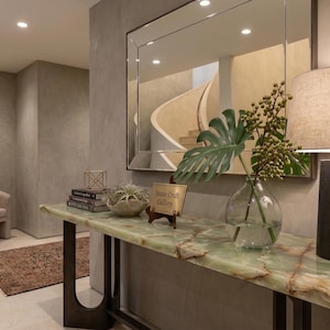May include: An interior shot of a hallway with a green marble console table and a black metal base. A lamp with a beige shade sits on the table, next to a glass vase with greenery. A mirror reflects a curved staircase.
