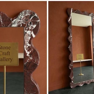 May include: A rectangular mirror with a wavy, marble-patterned frame in shades of red, brown, and white. The mirror has a gold sign that reads "Stone Craft Gallery". The mirror is displayed against a brown wall.