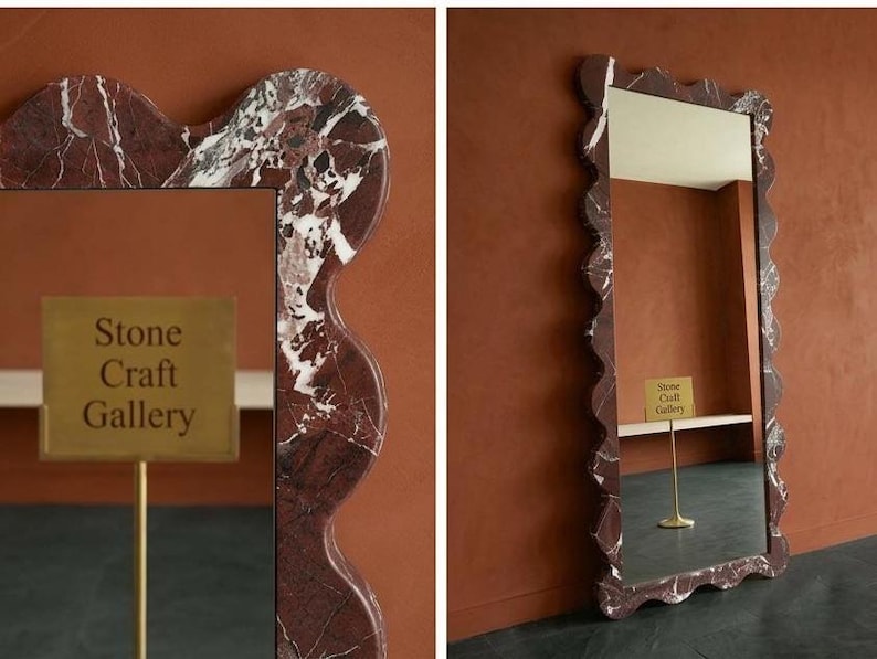 May include: A rectangular mirror with a wavy, marble-patterned frame in shades of red, brown, and white. The mirror has a gold sign that reads "Stone Craft Gallery". The mirror is displayed against a brown wall.