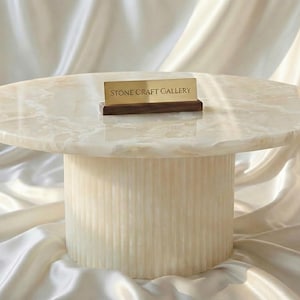 Pode incluir: Uma mesa de centro redonda, cor creme, com um tampo claro marmorizado e uma base cilíndrica. Uma pequena placa com a inscrição "STONE CRAFT GALLERY" está colocada em cima da mesa. O conjunto é apresentado sobre um fundo de tecido drapeado.