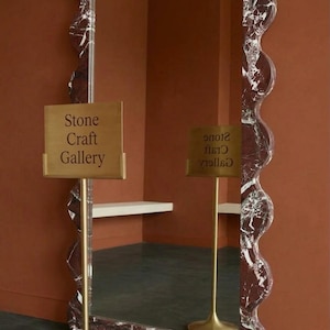 May include: A large, wavy-edged mirror with a red and white marble pattern. The mirror is displayed in a room with a sign that reads "Stone Craft Gallery". The sign is on a gold stand.