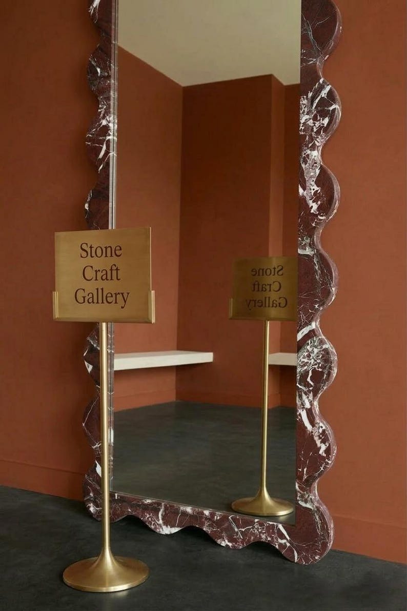 May include: A large, wavy-edged mirror with a red and white marble pattern. The mirror is displayed in a room with a sign that reads "Stone Craft Gallery". The sign is on a gold stand.