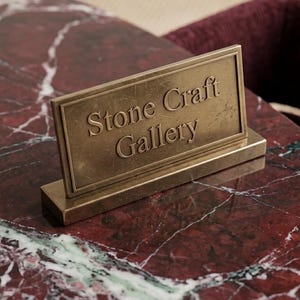 May include: A rectangular brass sign with the words "Stone Craft Gallery" in a serif font. The sign is set on a matching rectangular base. The background is a red and white marble surface.