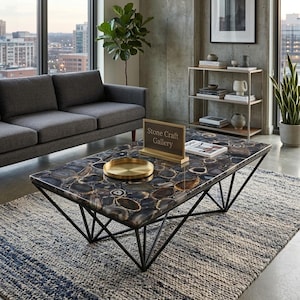May include: A rectangular coffee table with a dark, patterned stone top and a black metal geometric base. A gold-colored tray sits on the table. The rug is blue and white. The sign says "Stone Craft Gallery."