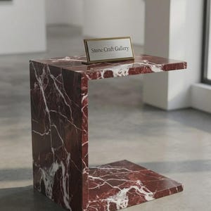 Puede incluir: Una mesa auxiliar en forma de C hecha de mármol rojo intenso con vetas blancas prominentes. Sobre la superficie superior hay un letrero metálico rectangular con marco dorado que muestra el texto Stone Craft Gallery impreso en negro sobre fondo plateado.