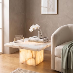 May include: A modern, illuminated onyx coffee table with a rectangular top and a square base. The table is in a living room setting, with a framed abstract art print, a cream-colored sofa, and a green throw blanket. A glass vase with white orchids sits on the table.