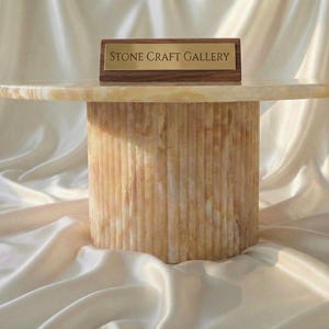 May include: A round, beige stone table with a cylindrical base. The table has a smooth, polished top and a textured, vertically-lined base. A small wooden sign with the text "STONE CRAFT GALLERY" is on top of the table.