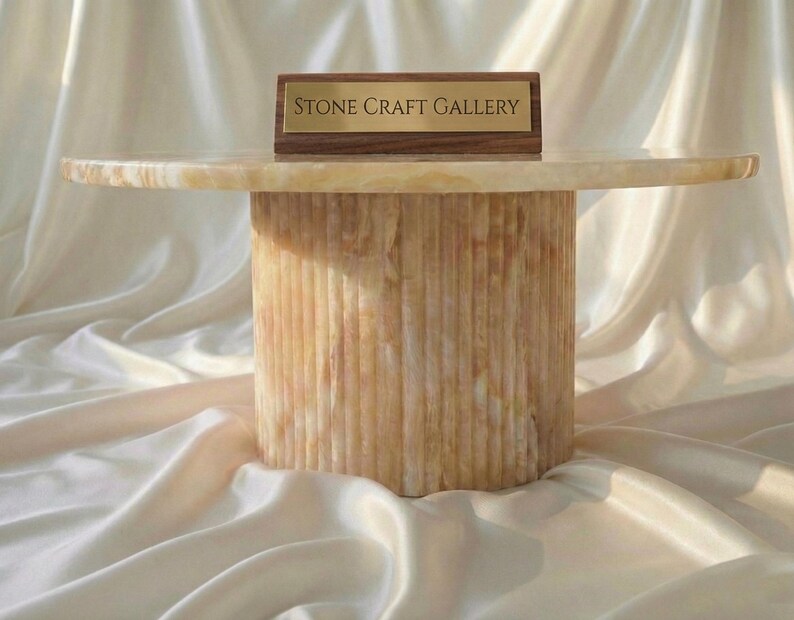 May include: A round, beige stone table with a cylindrical base. The table has a smooth, polished top and a textured, vertically-lined base. A small wooden sign with the text "STONE CRAFT GALLERY" is on top of the table.