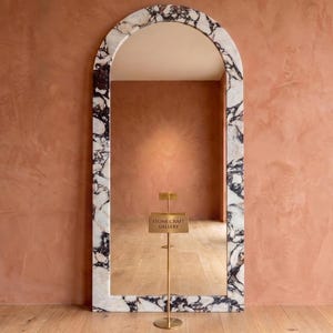May include: A tall, arched floor mirror featuring a thick frame made of white marble with prominent black veining. A thin, gold-toned metal stand with a small rectangular sign reading "STONE CRAFT GALLERY" is positioned directly in front of the mirror.