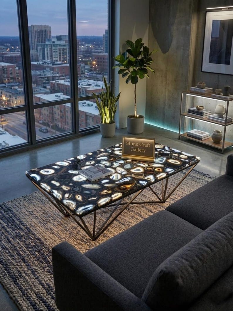 May include: A modern coffee table with a black and white stone top and a geometric metal base. The table is in a living room setting with a city view. The text on the table reads "Stone Craft Gallery".