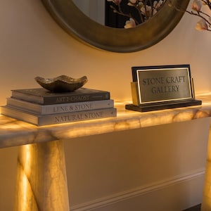 May include: A rectangular console table made of translucent, backlit stone with two tapered cylindrical legs. On top sits a stack of three books, a small dark bowl, a black sign with the gold text STONE CRAFT GALLERY, and a vase with flowers.