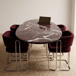 May include: An oval marble dining table with a deep red and white veined pattern, surrounded by four burgundy upholstered chairs with chrome frames. A small sign on the table reads "Stone Craft Gallery". The table and chairs are on a neutral-toned woven rug.