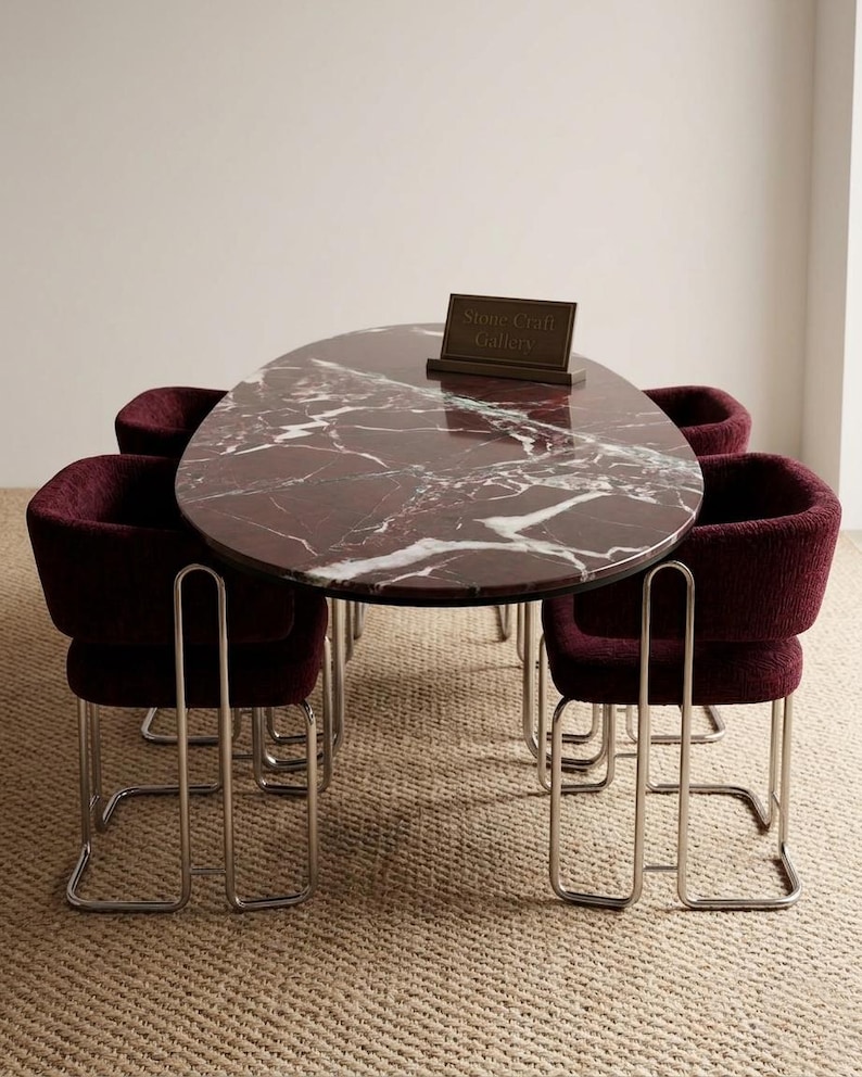 May include: An oval marble dining table with a deep red and white veined pattern, surrounded by four burgundy upholstered chairs with chrome frames. A small sign on the table reads "Stone Craft Gallery". The table and chairs are on a neutral-toned woven rug.