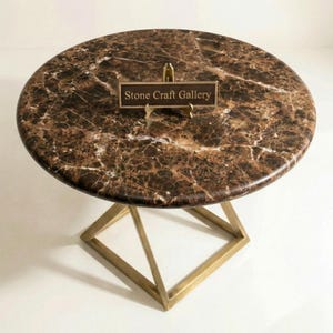 Könnte beinhalten: Runder Tisch mit einer Tischplatte aus braun-weißem Marmor. Das Gestell ist goldfarben und geometrisch. Ein kleines Schild auf dem Tisch trägt die Aufschrift "Stone Craft Gallery". Der Tisch hat einen Durchmesser von ca. 76 cm.