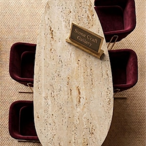 Pode incluir: Vista aérea de uma mesa oval de pedra com superfície texturizada, rodeada por três cadeiras bordô com estrutura dourada. Uma pequena placa na mesa diz "Stone Craft Gallery". A mesa tem cerca de 200 cm de comprimento.