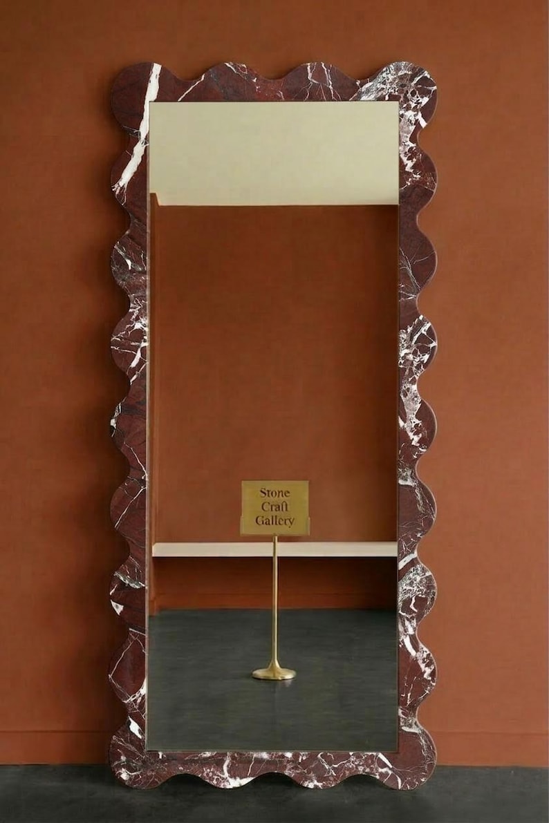 May include: A rectangular mirror with a wavy, red marble frame. The frame has white veining. The mirror reflects a dark floor and a sign that reads "Stone Craft Gallery". The background is a solid, warm brown.