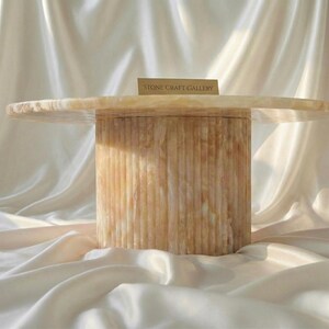 May include: A round, light-colored stone table with a cylindrical base. The base has vertical grooves, and the tabletop is smooth. The text "STONE CRAFT GALLERY" is on a small card on the table.