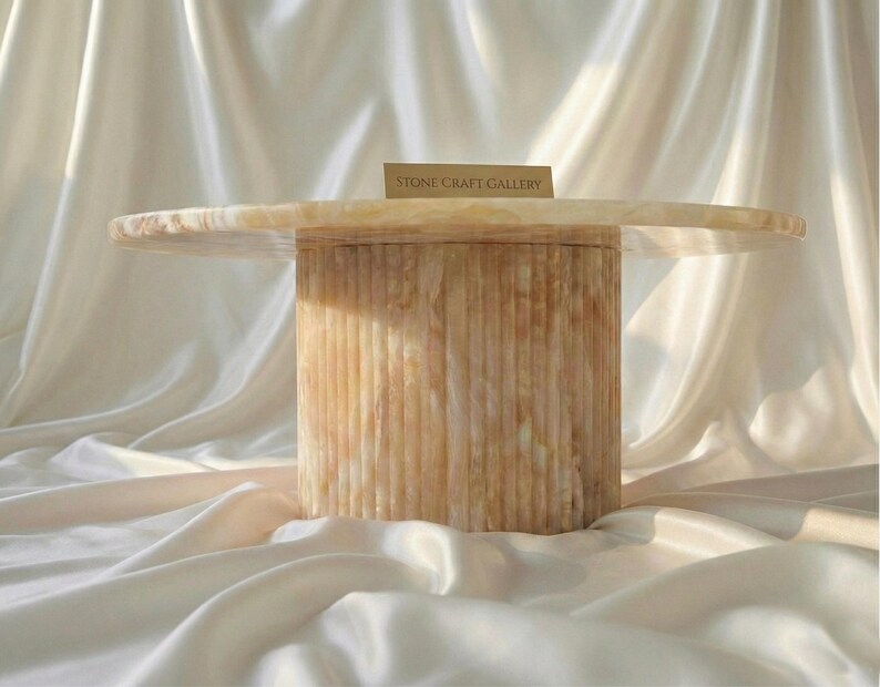 May include: A round, light-colored stone table with a cylindrical base. The base has vertical grooves, and the tabletop is smooth. The text "STONE CRAFT GALLERY" is on a small card on the table.