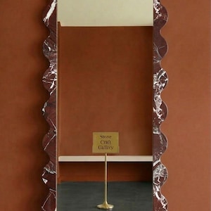 May include: A rectangular mirror with a wavy, red marble frame. The frame has white veining. The mirror reflects a dark floor and a sign that reads "Stone Craft Gallery". The background is a solid, warm brown.