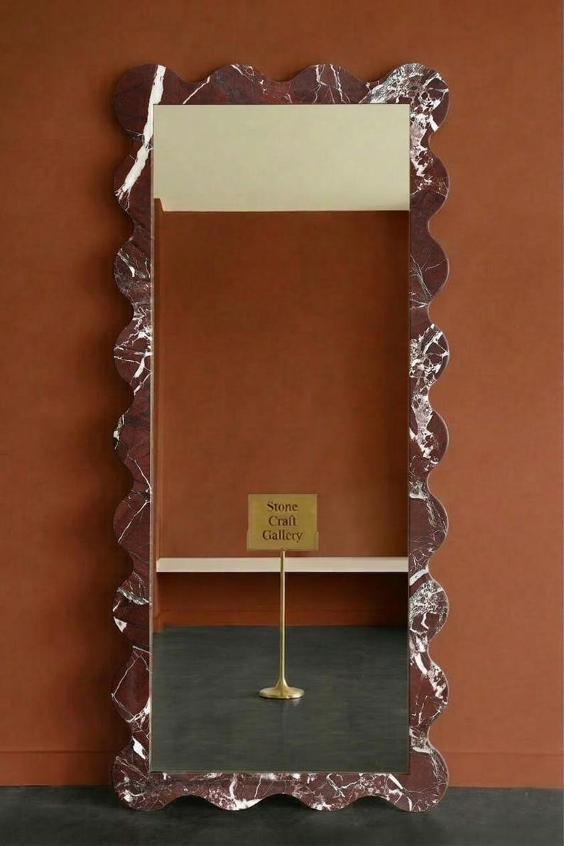 May include: A rectangular mirror with a wavy, red marble frame. The frame has white veining. The mirror reflects a dark floor and a sign that reads "Stone Craft Gallery". The background is a solid, warm brown.