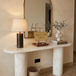 Puede incluir: Una consola de mármol con base cilíndrica y tapa ovalada. Una lámpara de color bronce con pantalla de lino se encuentra a la izquierda. Un espejo cuelga sobre la mesa y un letrero que dice "Stone Craft Gallery" está sobre la mesa.