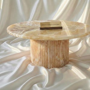 May include: A round, light-colored onyx coffee table with a cylindrical base. The tabletop has a marbled appearance, and the base features vertical grooves. A small wooden sign with the text "STONE CRAFT GALLERY" rests on the table.