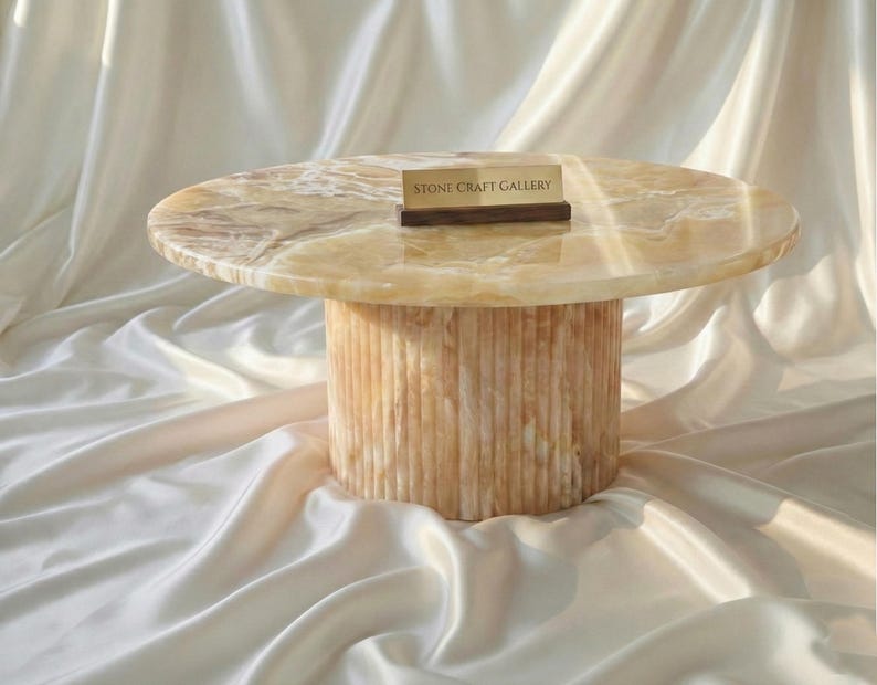 May include: A round, light-colored onyx coffee table with a cylindrical base. The tabletop has a marbled appearance, and the base features vertical grooves. A small wooden sign with the text "STONE CRAFT GALLERY" rests on the table.