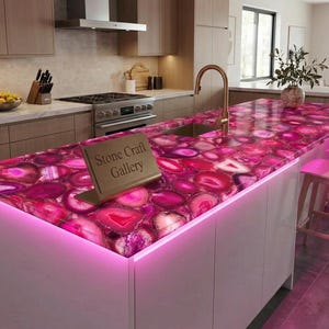 May include: A kitchen island with a vibrant pink agate countertop. The countertop features a polished surface with a unique pattern of pink, white, and purple hues. The island has a white base with pink under-lighting, a sink, and a gold faucet. A sign reads "Stone Craft Gallery."