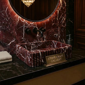 May include: A rectangular red marble sink with white veining, featuring a fluted exterior. The sink is set against a matching marble wall with a round mirror and black fixtures. A sign reads "STONE CRAFT GALLERY."