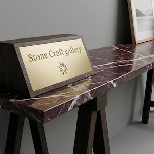 May include: A dark wood sign with a gold plaque that reads "Stone Craft gallery" and a star symbol. The sign sits on a long, marble-topped table with dark wood legs. A framed picture and a vase of flowers are also on the table.