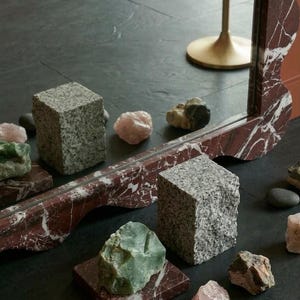 May include: An artistic display of natural stone elements. The image features a mirror with a red and white marble design, granite cubes, and various raw gemstones in shades of green and pink. The text "Stone Craft Gallery" is visible.