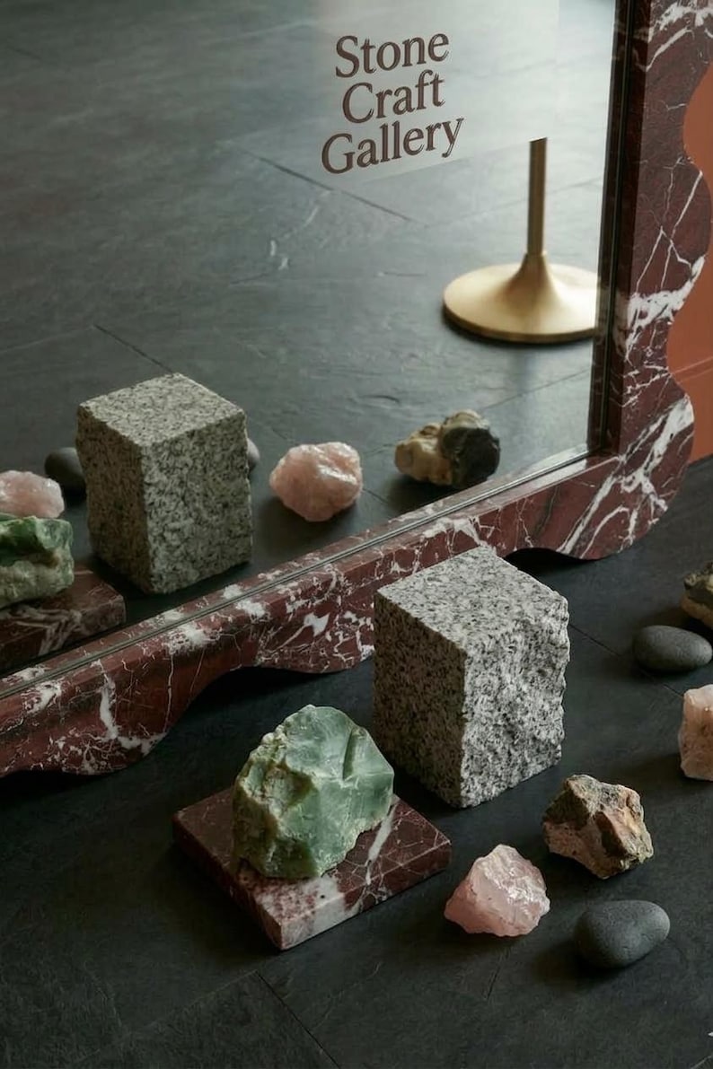 May include: An artistic display of natural stone elements. The image features a mirror with a red and white marble design, granite cubes, and various raw gemstones in shades of green and pink. The text "Stone Craft Gallery" is visible.