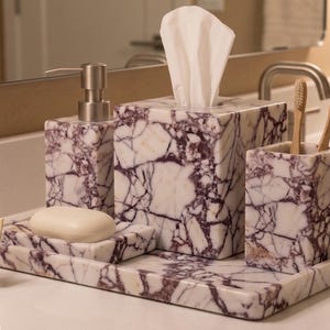 May include: A marble bathroom set with a soap dish, soap dispenser, tissue box, toothbrush holder, and tray. The set has a white and purple veined pattern. A small easel with the text "Stone Craft Gallery" is in the foreground.