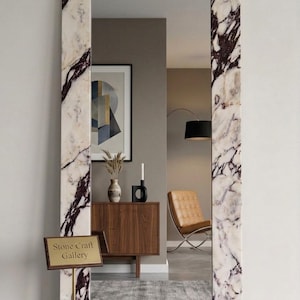 May include: Full-length rectangular mirror with a white marble frame featuring dark purple veining. The mirror reflects a room with a wooden cabinet, artwork, and a leather chair. A sign in front of the mirror reads "Stone Craft Gallery."