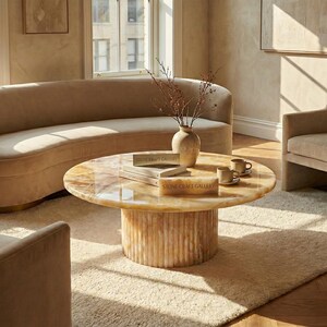 May include: A living room scene featuring a curved beige sofa, two armchairs, and a round coffee table. The coffee table has a light-colored stone top and a ribbed base. A beige rug and a vase with dried branches complete the decor.