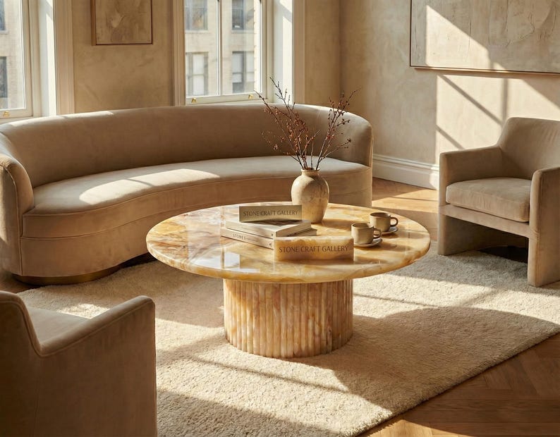 May include: A living room scene featuring a curved beige sofa, two armchairs, and a round coffee table. The coffee table has a light-colored stone top and a ribbed base. A beige rug and a vase with dried branches complete the decor.