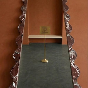 May include: A tall, rectangular floor mirror featuring a thick, wavy-edged frame made of dark red marble with white veining. The reflective surface shows a gold-colored stand with a sign that reads Stone Craft Gallery in the center.