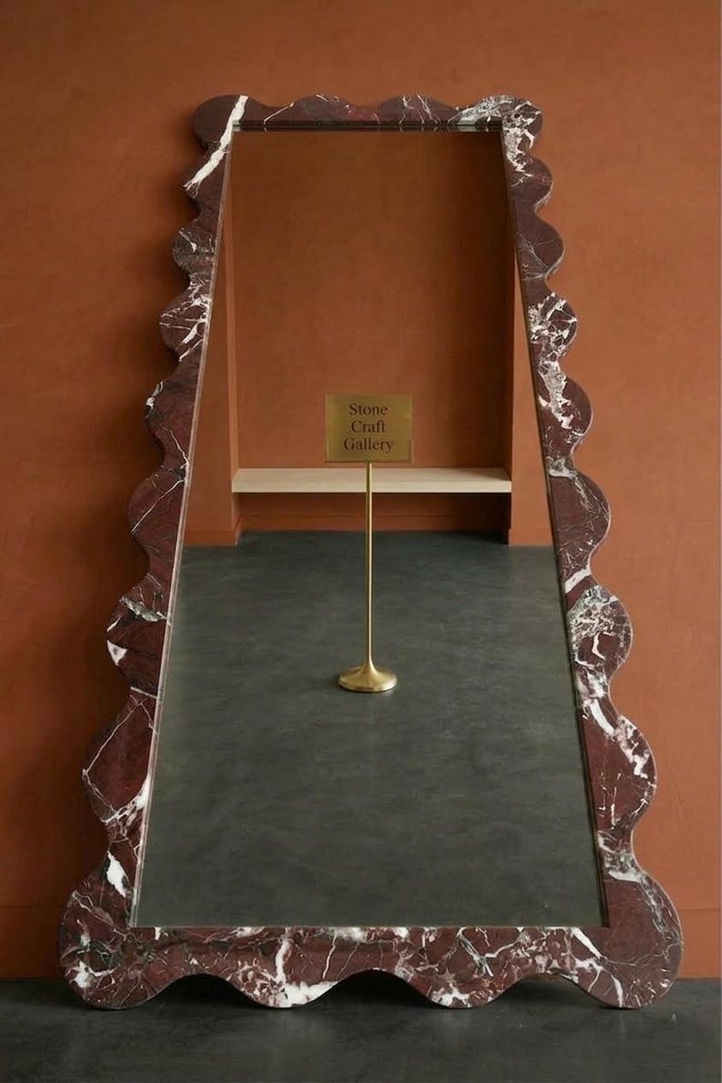 May include: A tall, rectangular floor mirror featuring a thick, wavy-edged frame made of dark red marble with white veining. The reflective surface shows a gold-colored stand with a sign that reads Stone Craft Gallery in the center.