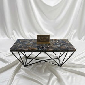 May include: A rectangular coffee table with a patterned stone top in shades of brown, blue, and gray. The table has a geometric black metal base. A small sign with the text "Stone Craft Gallery" is on the table.