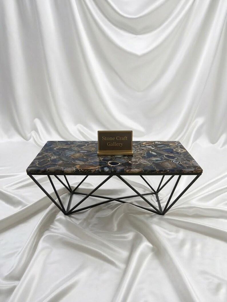 May include: A rectangular coffee table with a patterned stone top in shades of brown, blue, and gray. The table has a geometric black metal base. A small sign with the text "Stone Craft Gallery" is on the table.