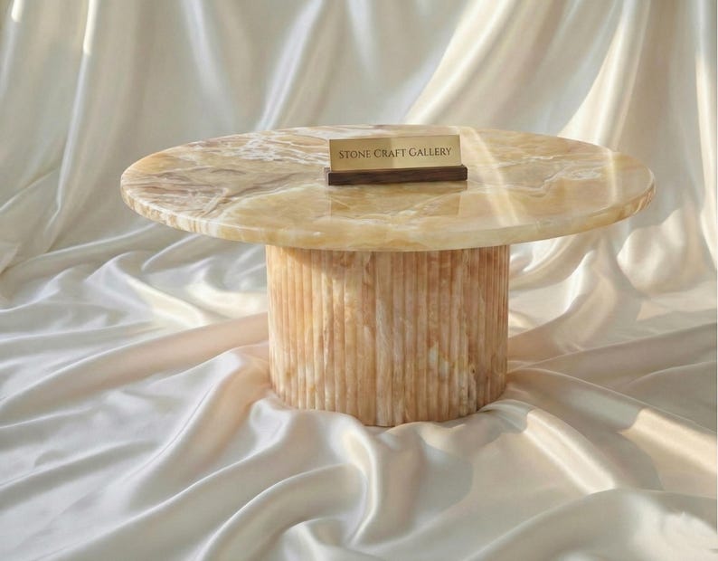 May include: A round, light-colored onyx coffee table with a cylindrical base. The tabletop has a marbled appearance, and the base features vertical grooves. A small wooden sign with text is on the table.