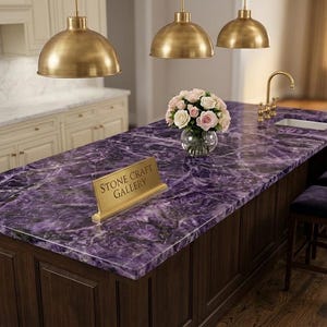Può includere: Un'isola cucina con un sorprendente piano di lavoro viola, con un motivo simile al marmo. L'isola ha mobili in legno scuro ed è adornata da un cartello dorato con la scritta "STONE CRAFT GALLERY". Lampade a sospensione dorate sono appese sopra e due sedie di velluto viola sono posizionate nelle vicinanze.