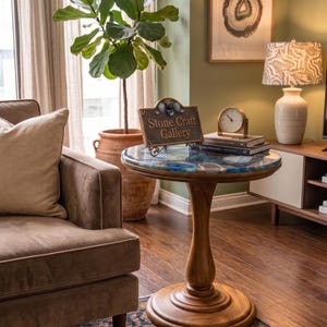 May include: A wooden side table with a blue and white resin top. The table has a turned pedestal base. The table is in a living room setting with a sofa, plant, and artwork. A sign on the table reads "Stone Craft Gallery."